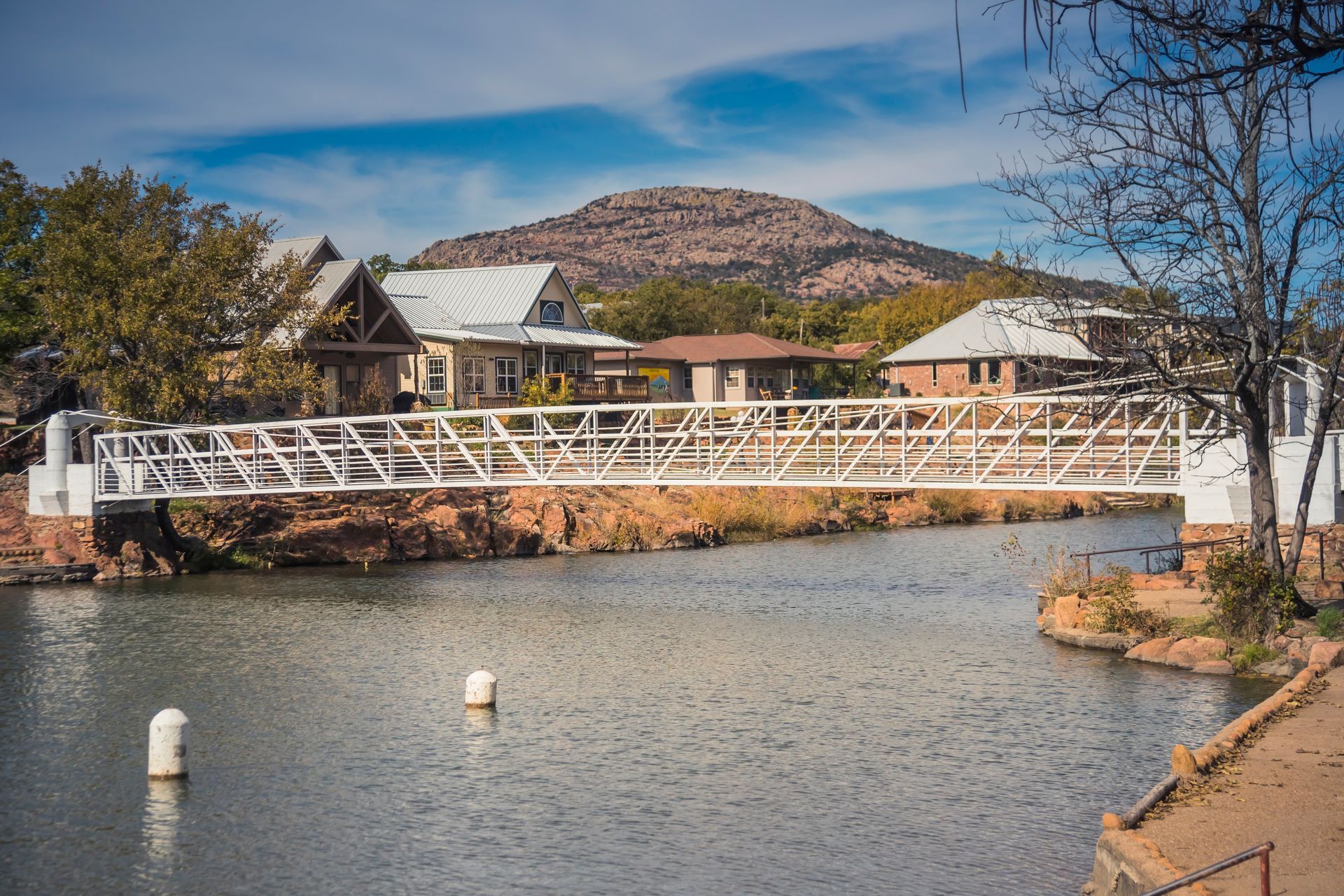 White pedestrian bridge over a river with houses and a rocky hill under a blue sky