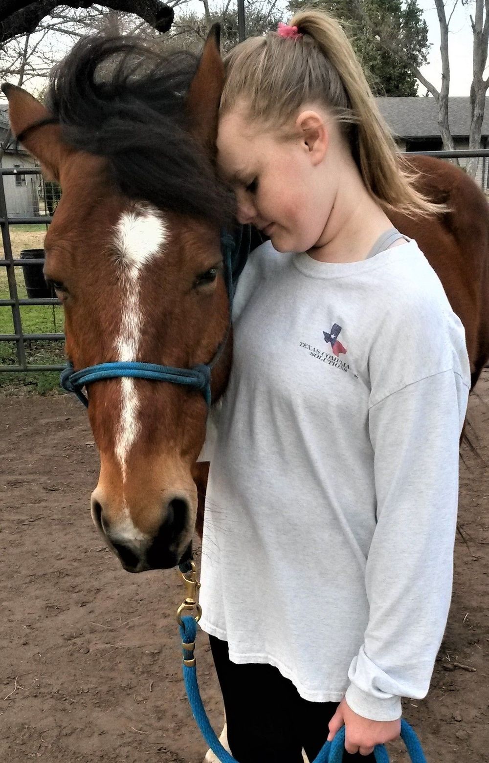 Girl with blonde ponytail hugs a brown horse with a white blaze. They are outdoors, possibly a stable.