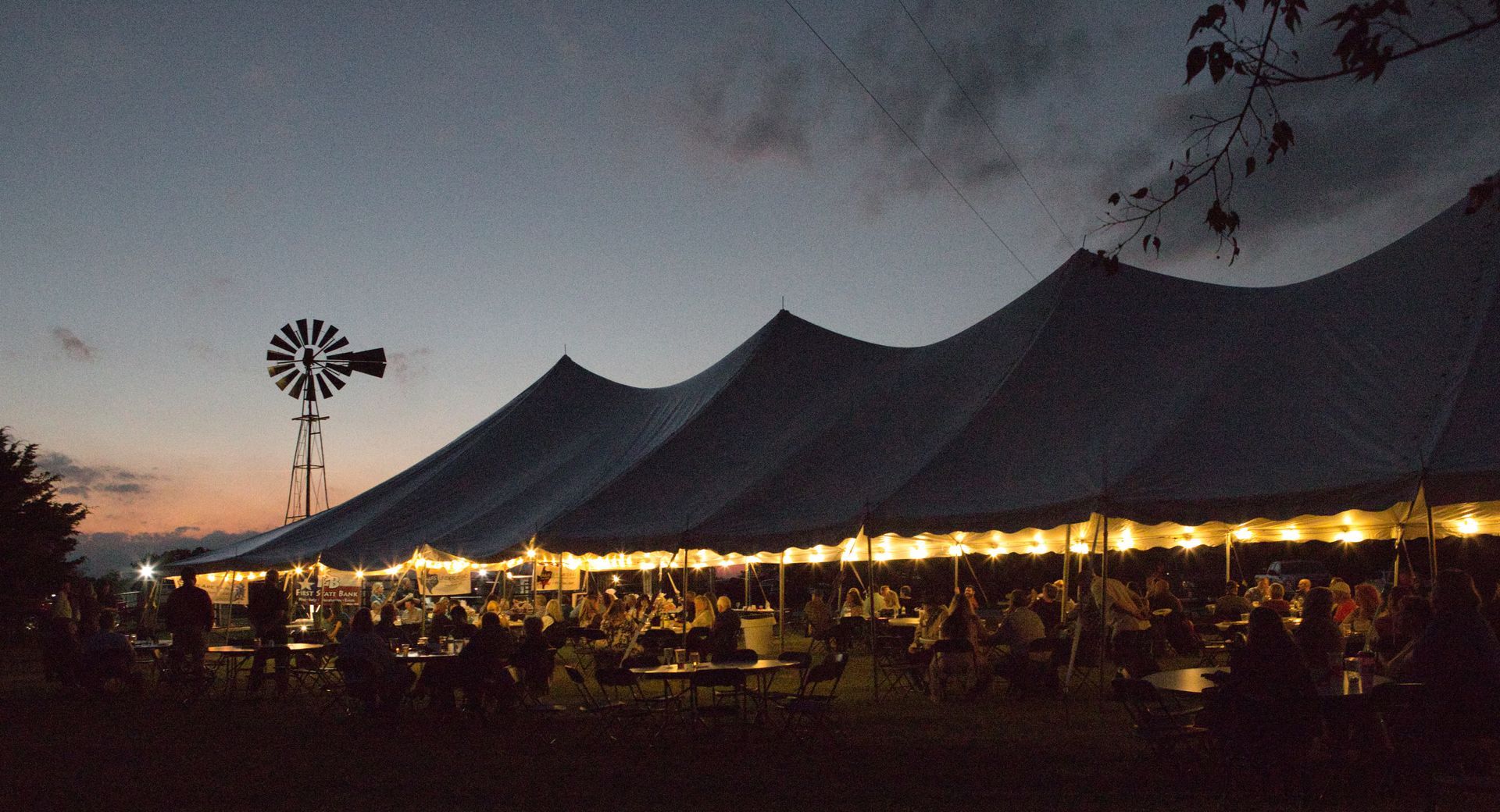 Large tent lit with string lights, people dining inside at dusk, windmill in background.