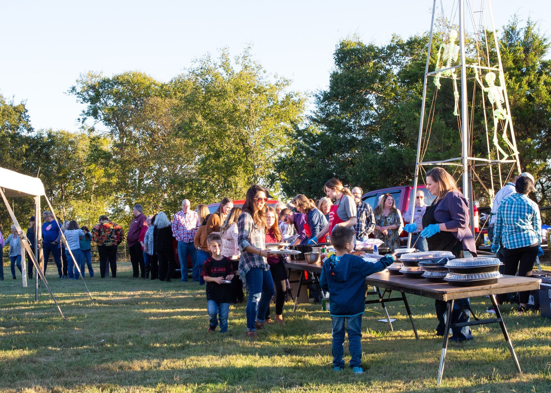 People line up for food at an outdoor event, with tables, tents, and a windmill in the background.