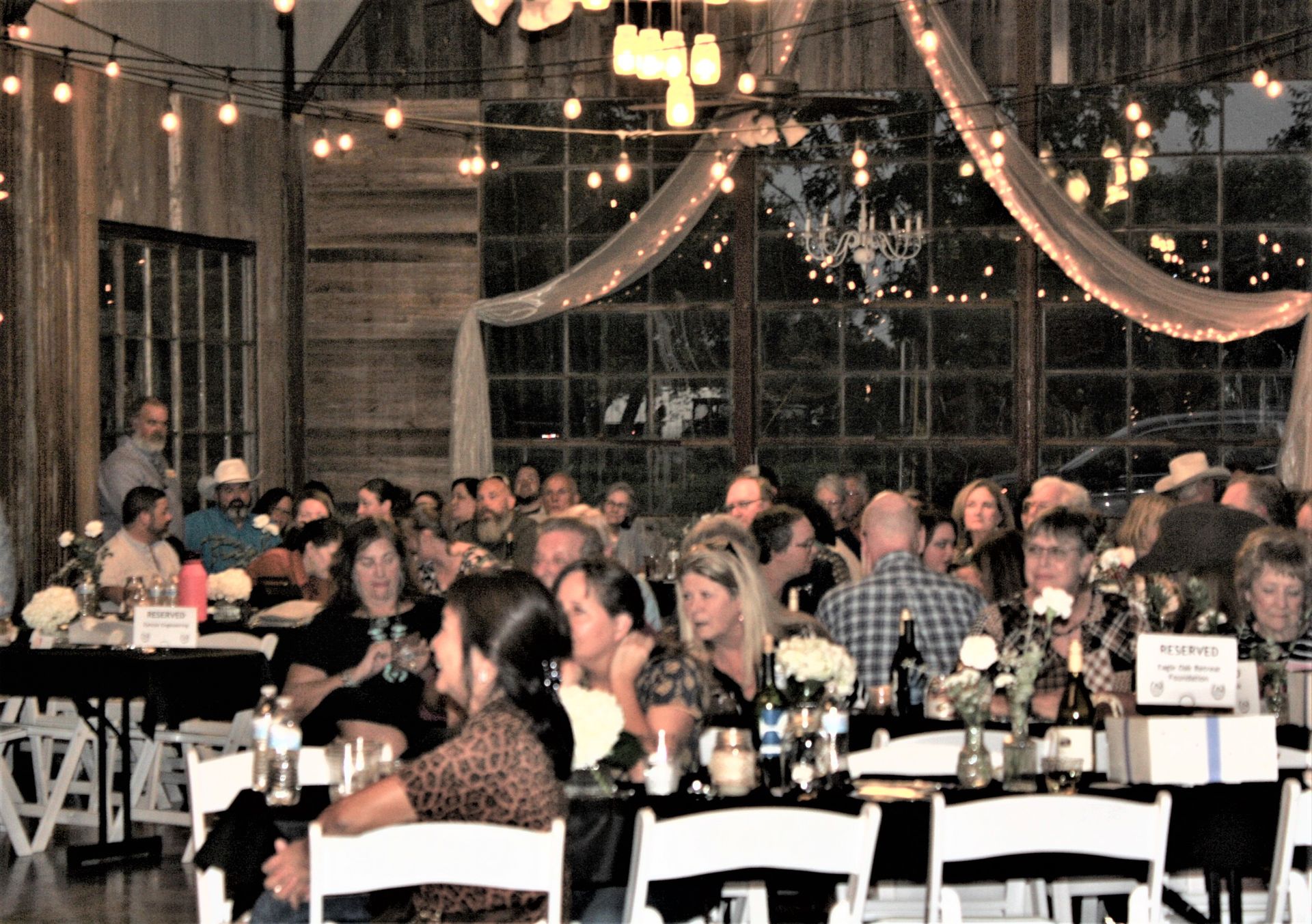 Large event in a barn with people seated at tables, lights strung, large windows.
