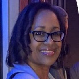 Woman with dark skin wearing glasses, smiling, in a dimly lit environment.