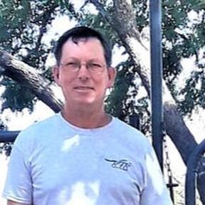 Man with glasses in light blue shirt smiles outdoors near trees.