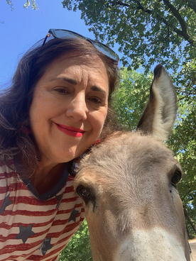 Woman smiles next to a donkey in front of trees on a sunny day.