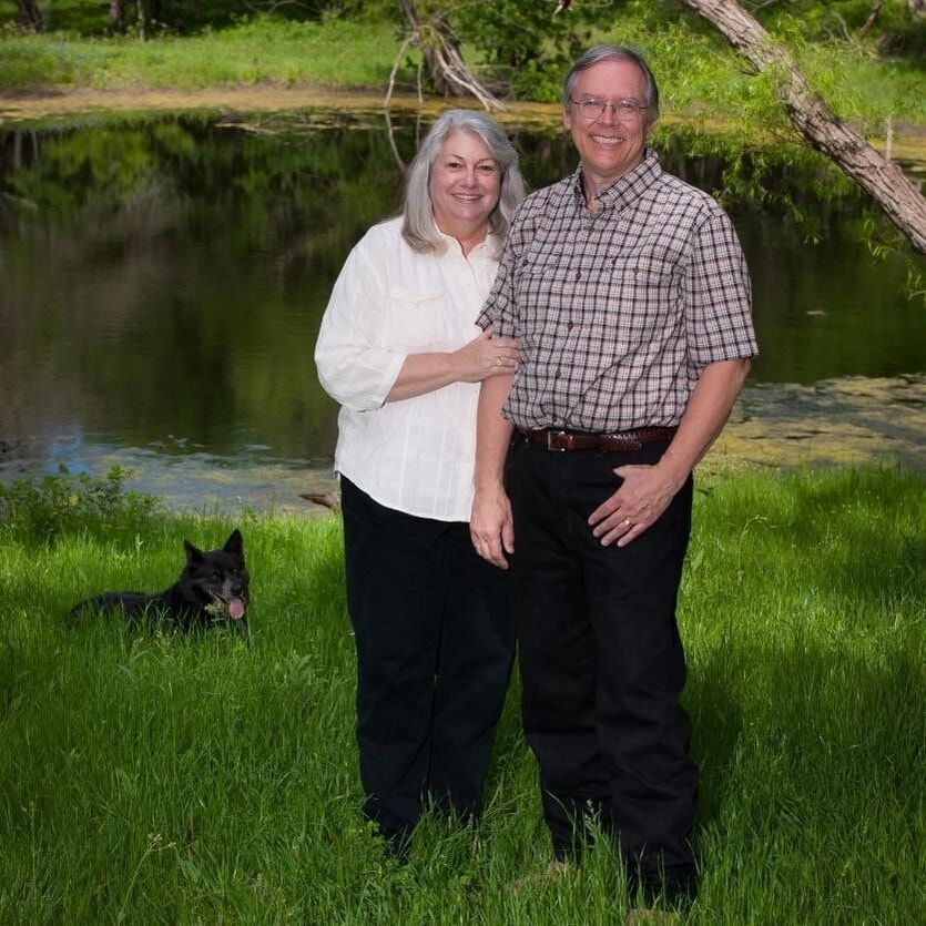 A couple stands by a pond in grass with a dog. Woman in white shirt, man in plaid shirt.