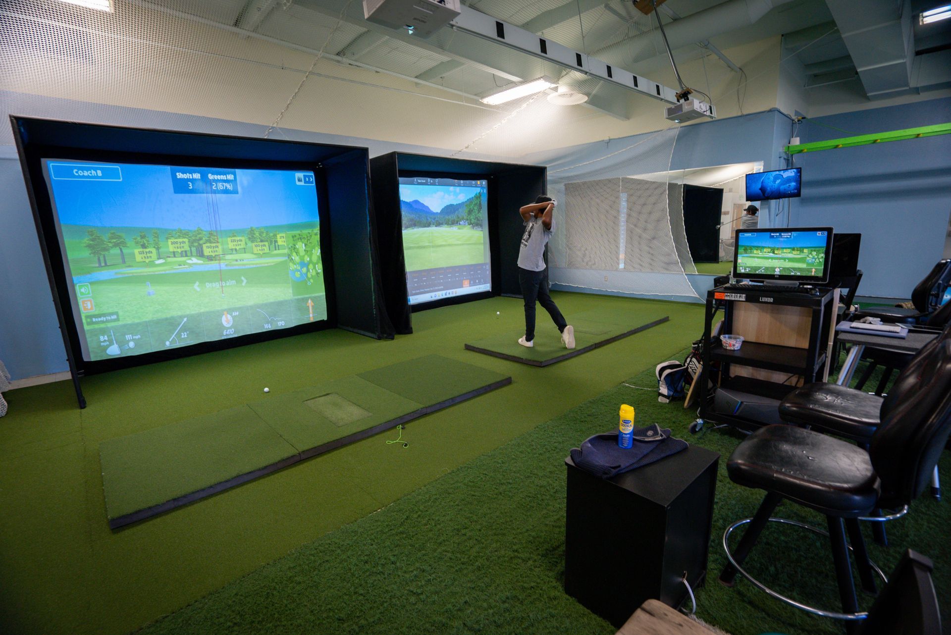 Indoor golf simulator: Woman swings club towards screen displaying golf course. Green carpet and dark room.