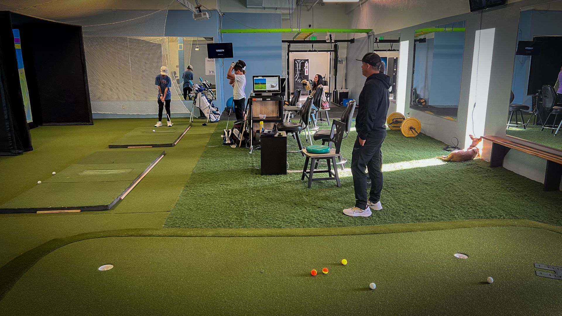 Golf training facility. People practicing swings on artificial turf. Several balls are scattered about.
