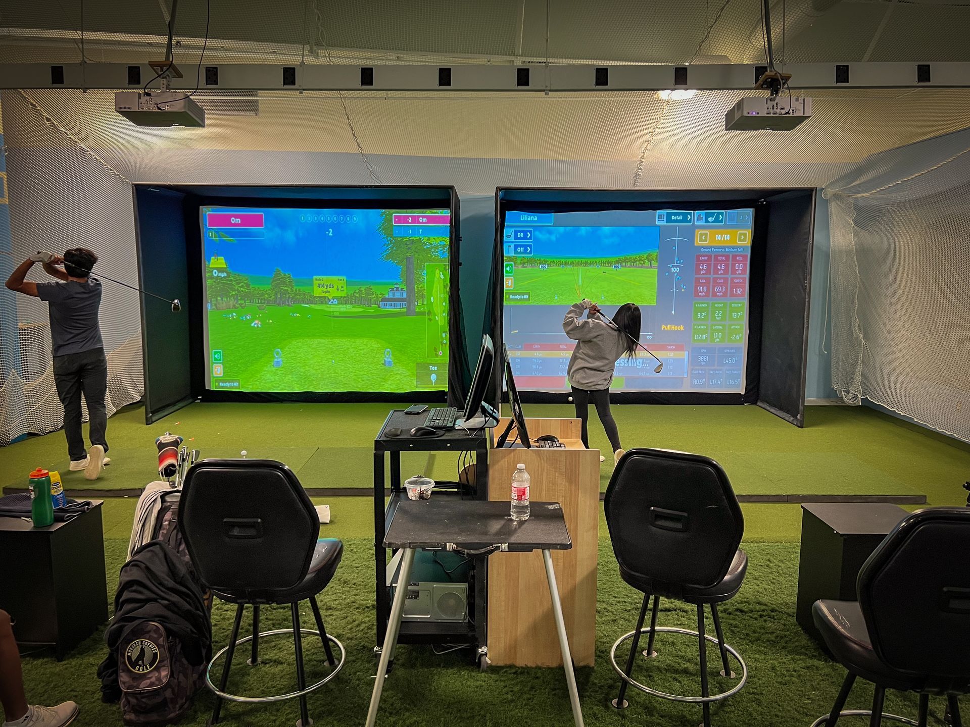 Two people playing golf in an indoor simulator, with screens displaying a golf course.