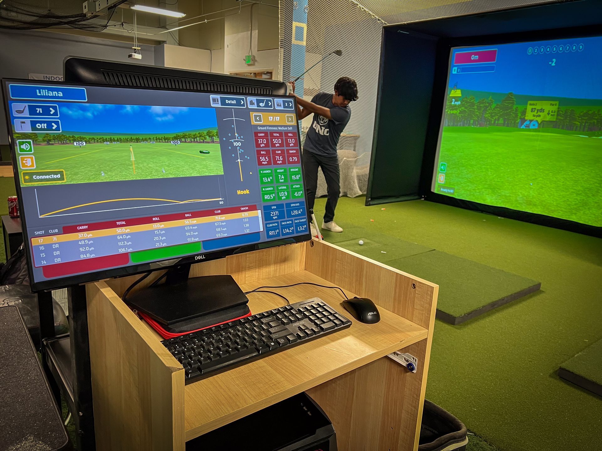 A man swings a golf club at a simulator. A large monitor displays data, with a keyboard and mouse on a wooden desk.