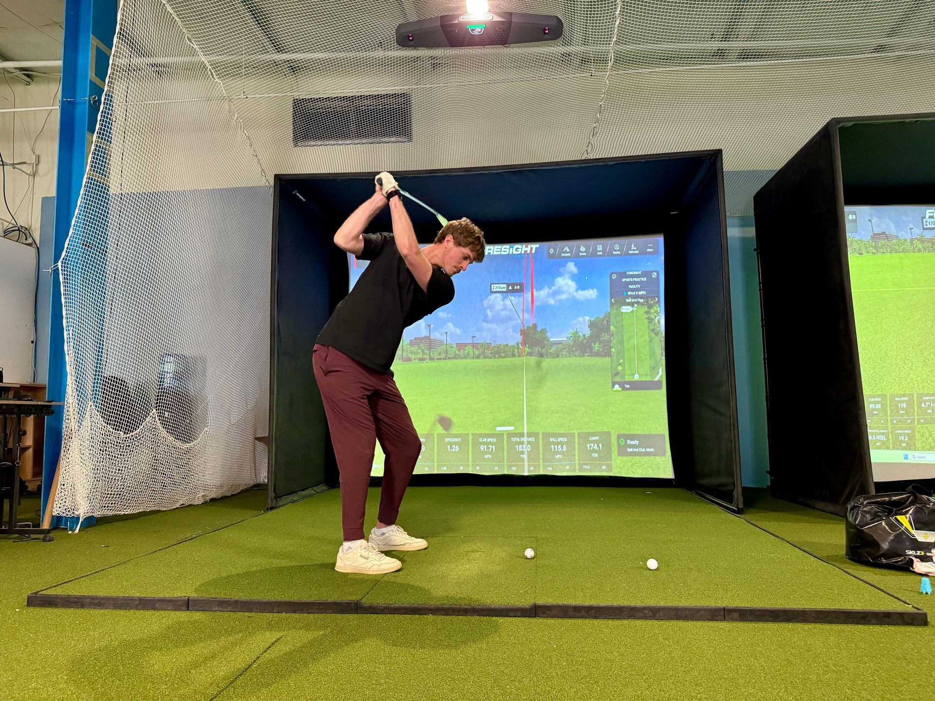 Man swinging a golf club in a golf simulator. He wears a black shirt, red pants, and white shoes.