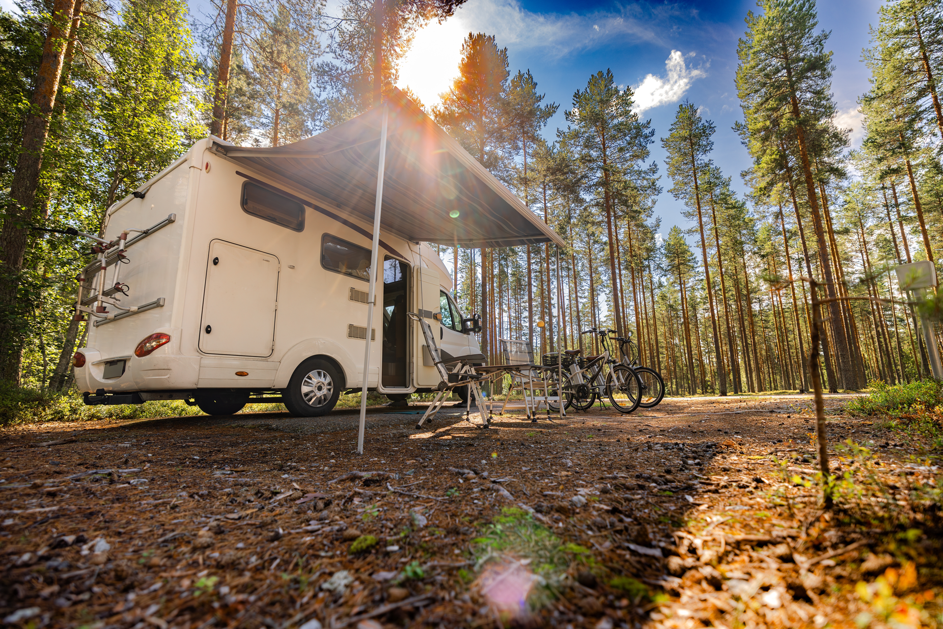 RV parked in a forest campsite with awning extended. Sunlight filters through the tall trees.