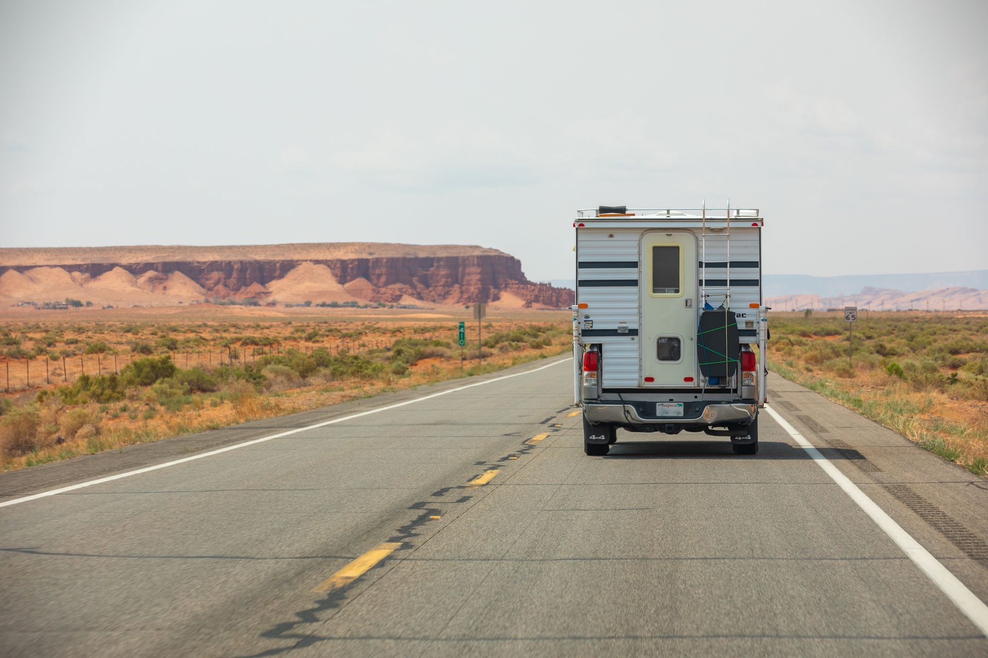 White camper truck driving on a desert highway with a mesa in the background.