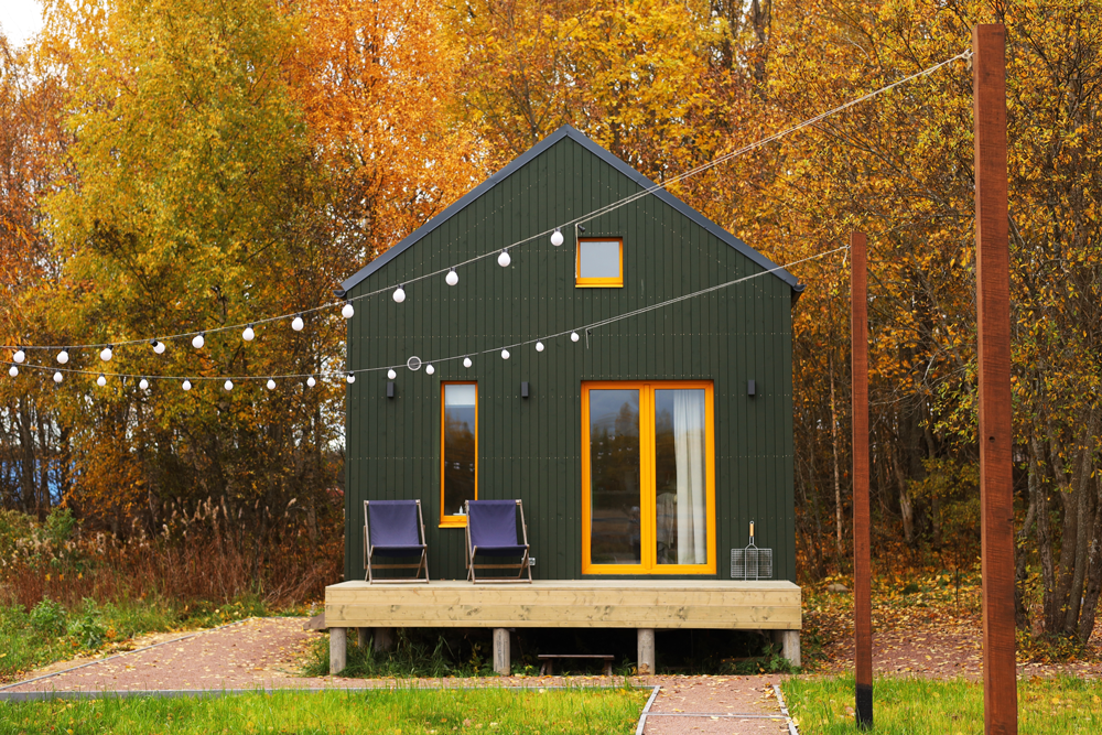 Dark green cabin with yellow door and windows, surrounded by fall foliage. String lights and two chairs on a wooden deck.