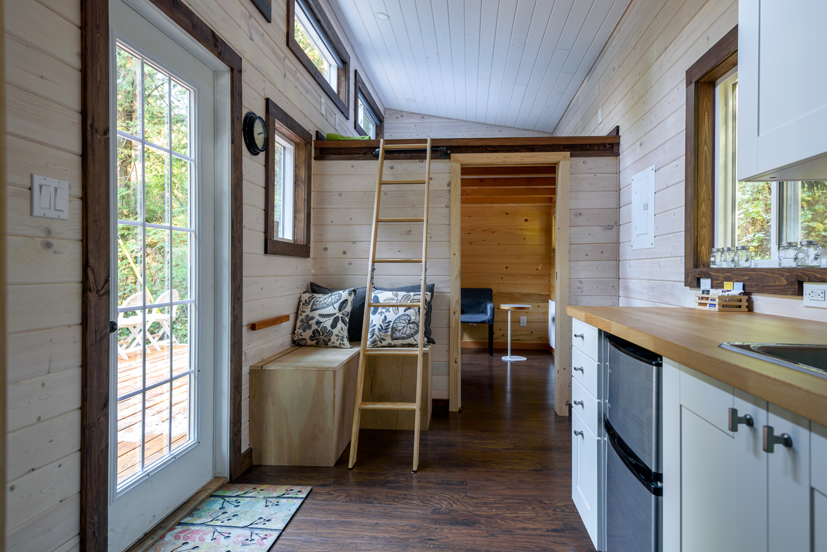 Interior of a tiny house with a bench, ladder to a loft, and kitchen counter with a window.