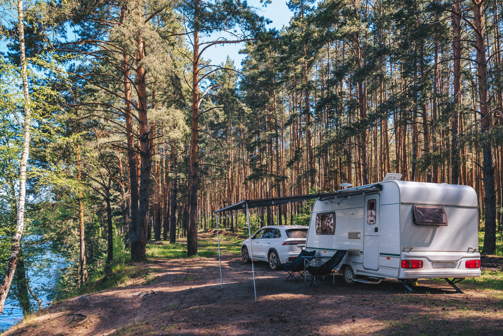 White camper parked near a car on a wooded campsite by water, awning extended.