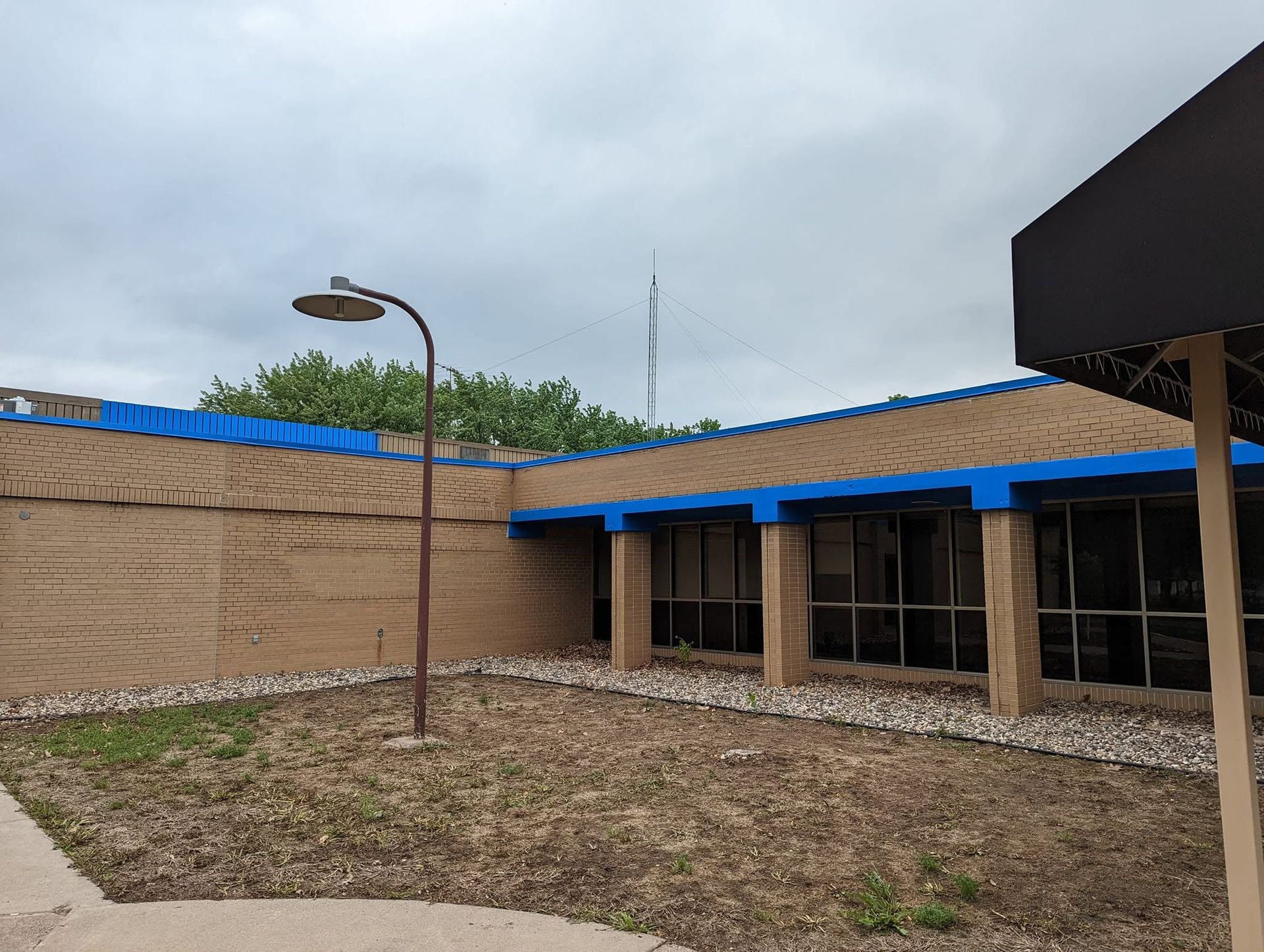 Empty courtyard with a beige building, blue roof trim, and a lamppost under a cloudy sky.