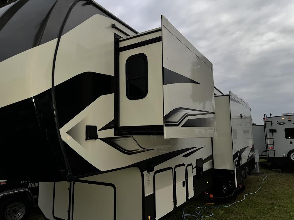 Side view of a large RV with beige and black paint, slide-out extended, outdoors on a cloudy day.