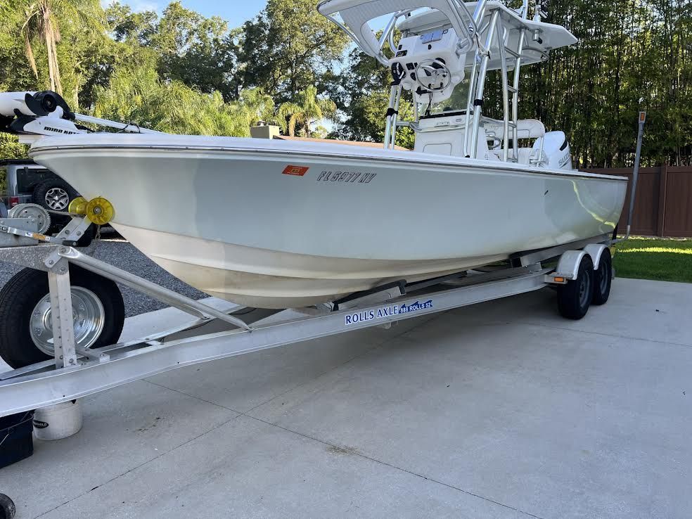 White fishing boat on a trailer, parked on grass. Trees and sky in the background.