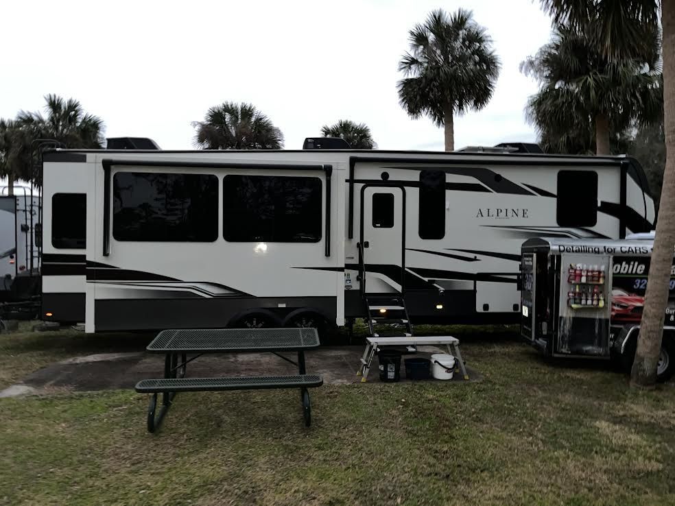 Large white and black RV parked on grass, picnic table in front. Trailer attached.