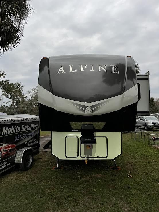 Front view of a black and white Alpine fifth-wheel camper. It's parked on grass, with a detailing trailer to the left.