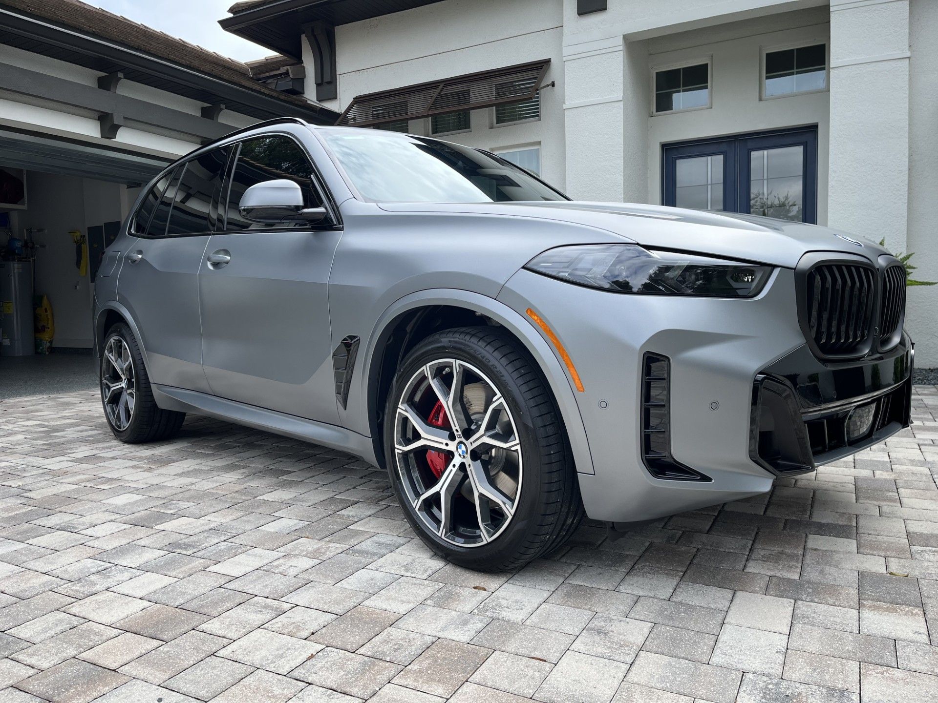Gray BMW X5 SUV parked on a brick driveway, red brake calipers, black grille, in front of a house.