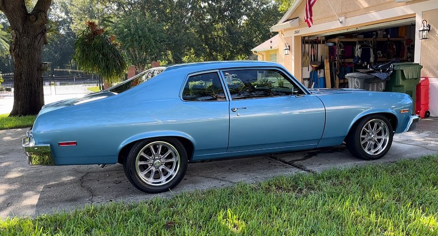 Light blue classic car parked in front of a house with a garage; sunny day.