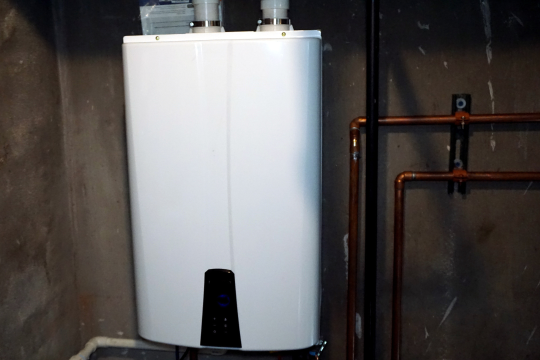 A white water heater is sitting in a dark room next to copper pipes.