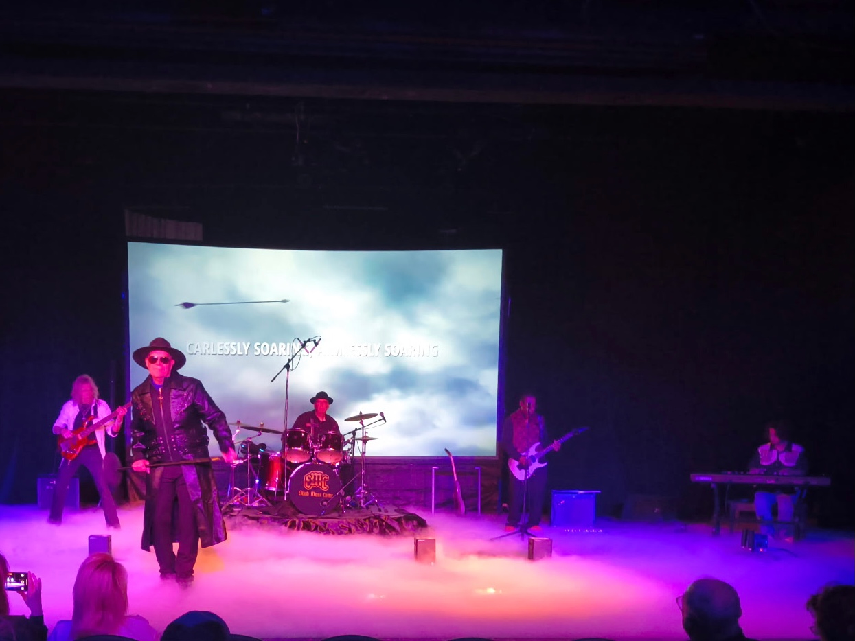 Band performing on stage with a backdrop; lead singer in coat, musicians playing instruments, stage smoke.