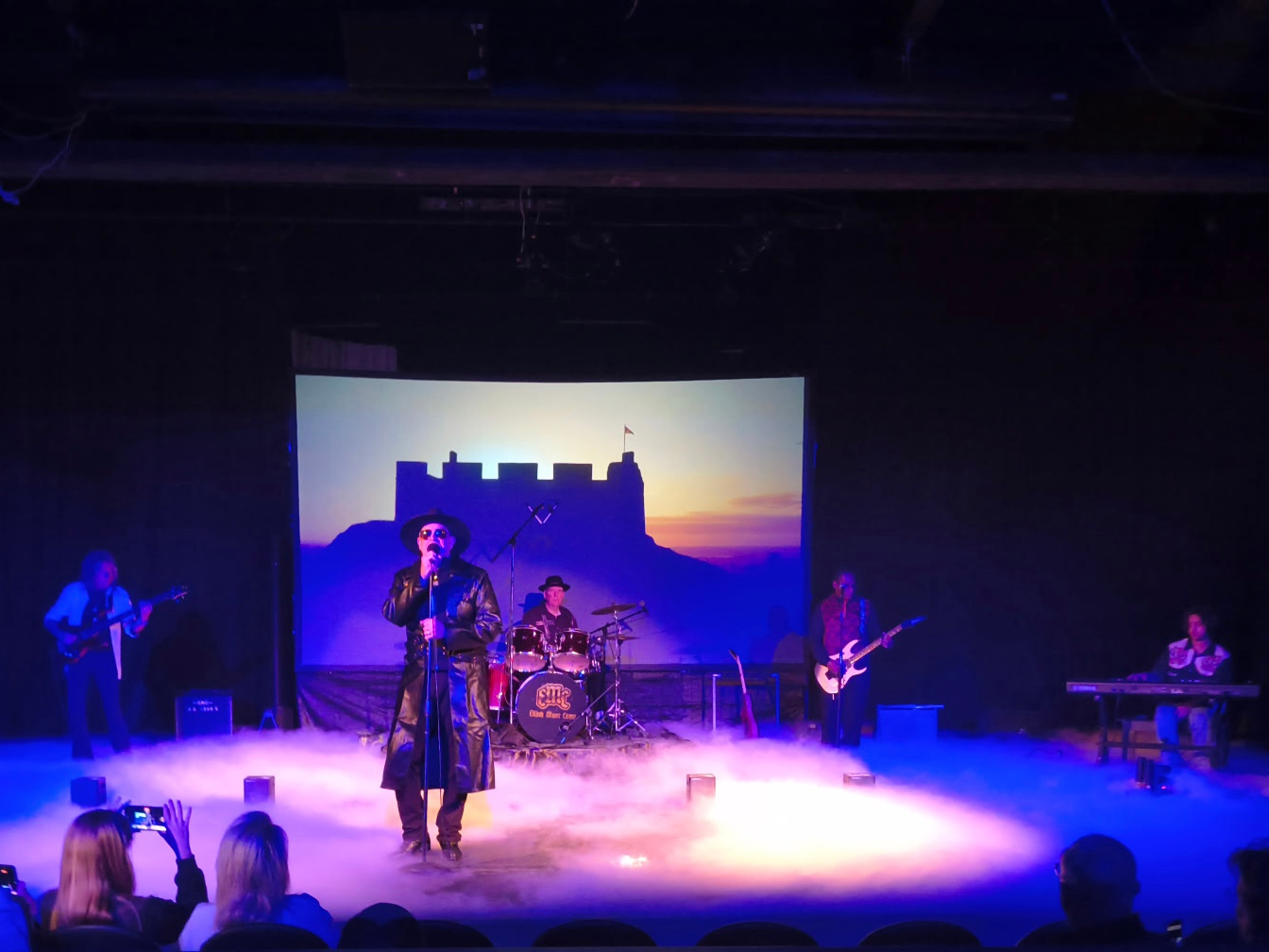 Band performing on stage with a castle backdrop; singer in a long coat, musicians with instruments, and audience visible.