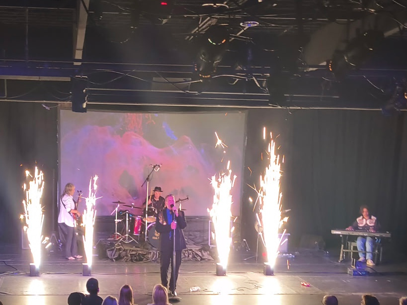 Band on stage with pyrotechnics. Singer with microphone, drummer, keyboardist, and background screen.