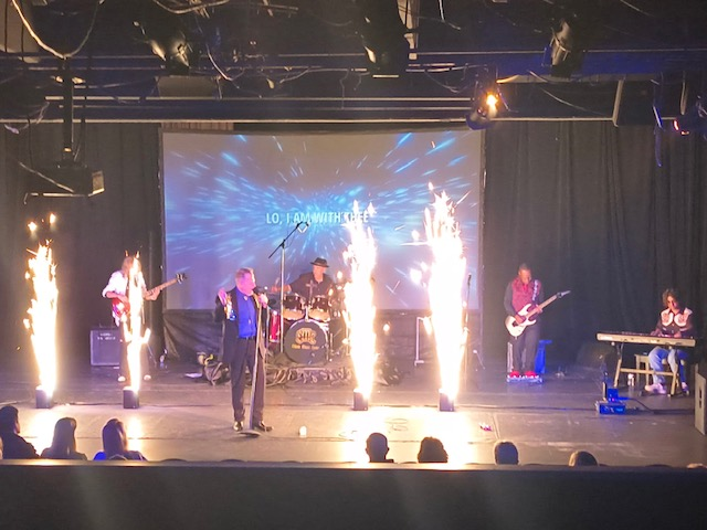 Band performing on stage with pyrotechnics. Screen in background. Audience in the foreground.