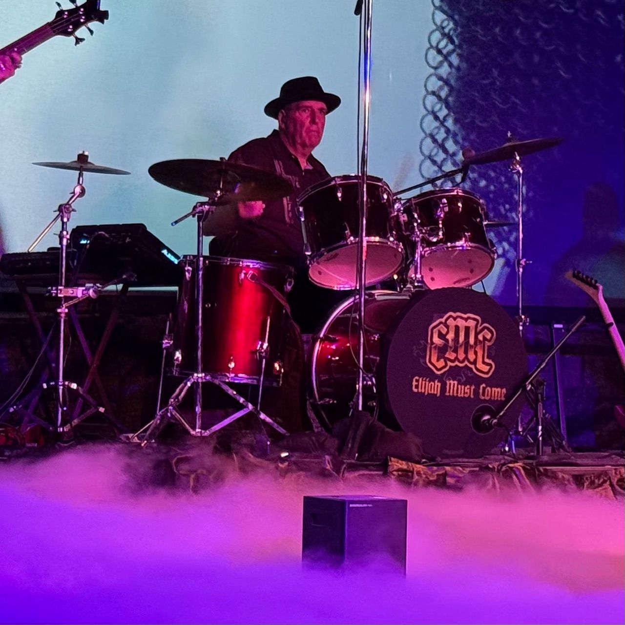 Drummer playing a black drum kit on stage, wearing a black hat. Stage has purple and white fog.
