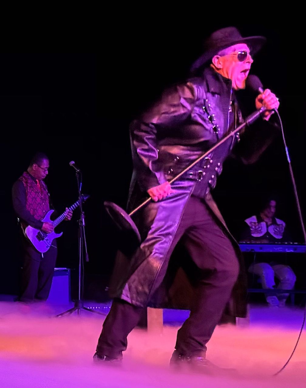 Singer on stage in black leather coat, singing into a microphone. Guitarist and keyboardist in background.