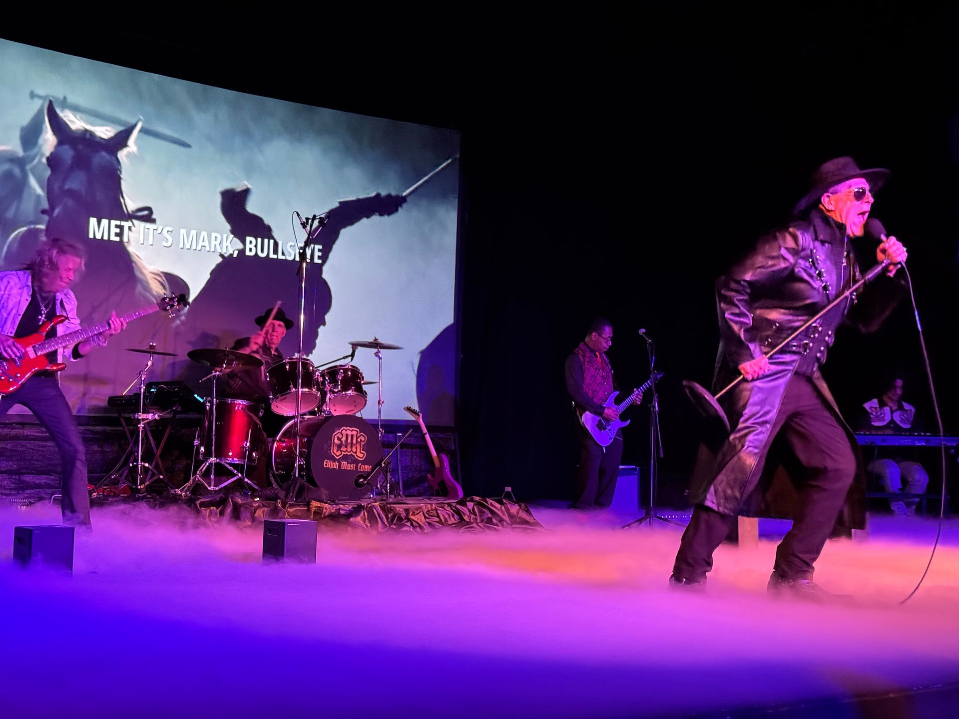 A band performing on stage, lead singer with a mic, guitarist, drummer.  Smoke and a screen with a fantasy image.