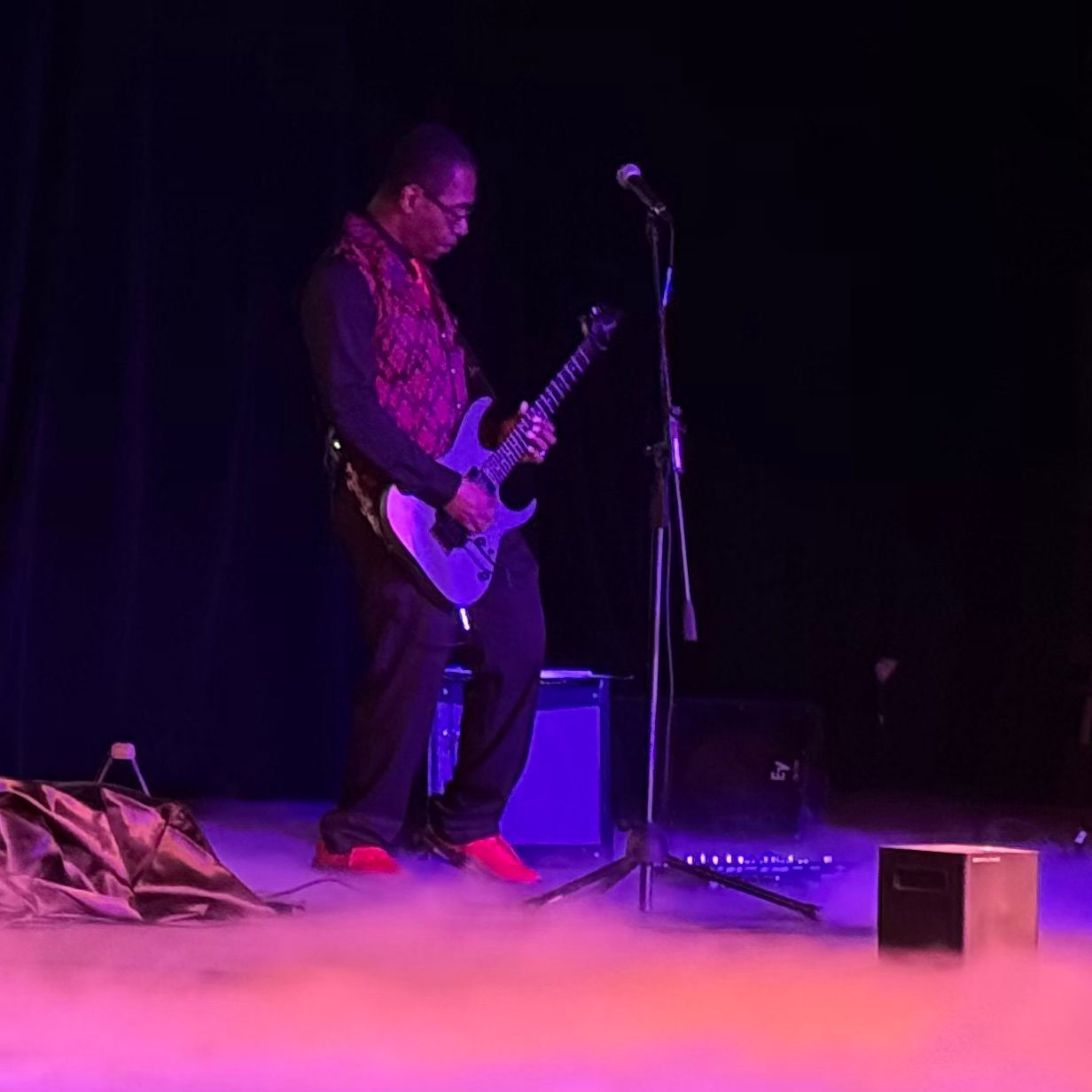 Man playing electric guitar onstage with purple lighting and stage fog.
