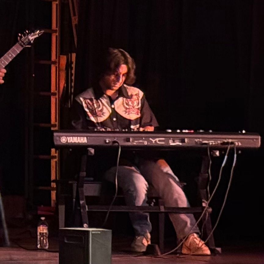 Person playing a Yamaha keyboard on stage, wearing a patterned shirt and jeans. Dark setting.
