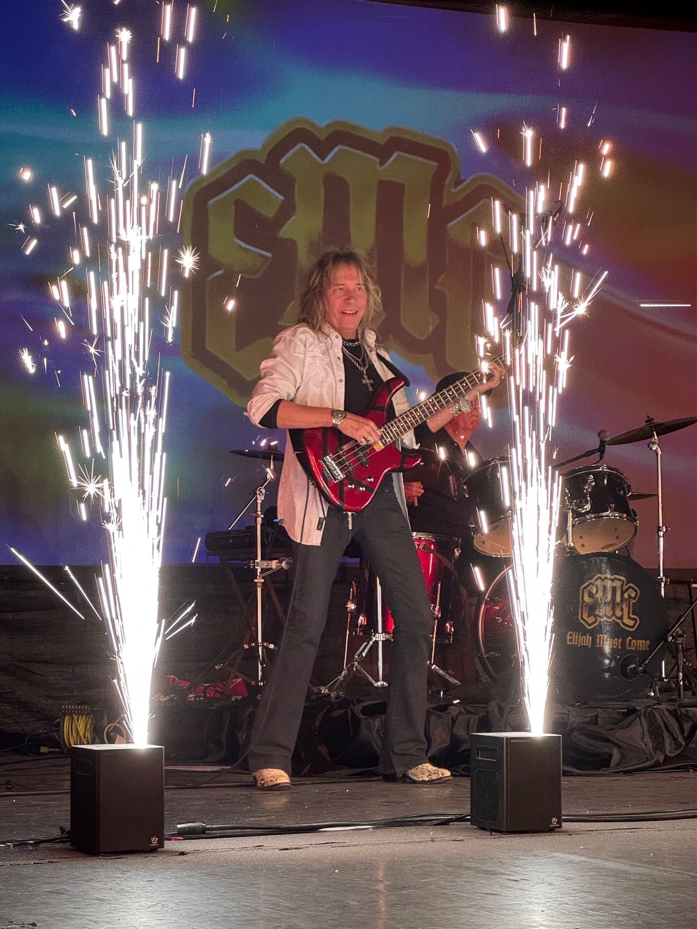 Guitarist on stage with fireworks, band logo behind, red guitar, stage lights.