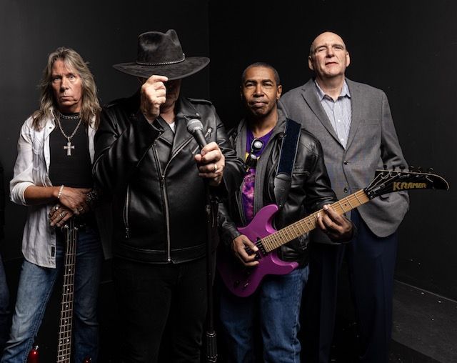 Rock band posing against a dark background: singer with a black hat and jacket, guitarist with a purple guitar, and two other band members.