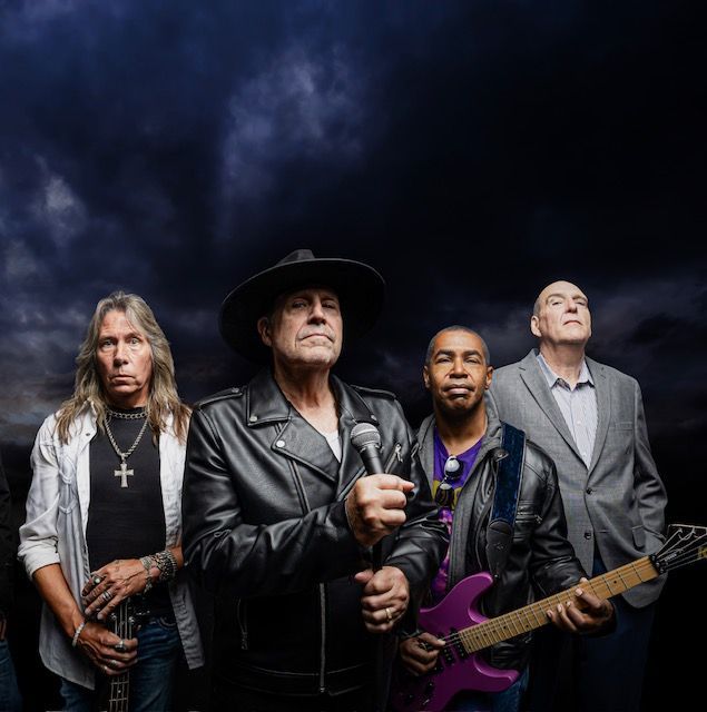Band of five men posing against a dark sky; one holding a microphone, one with a guitar.