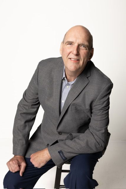 Man in gray blazer, blue shirt, dark pants, seated on stool, smiling, against white backdrop.