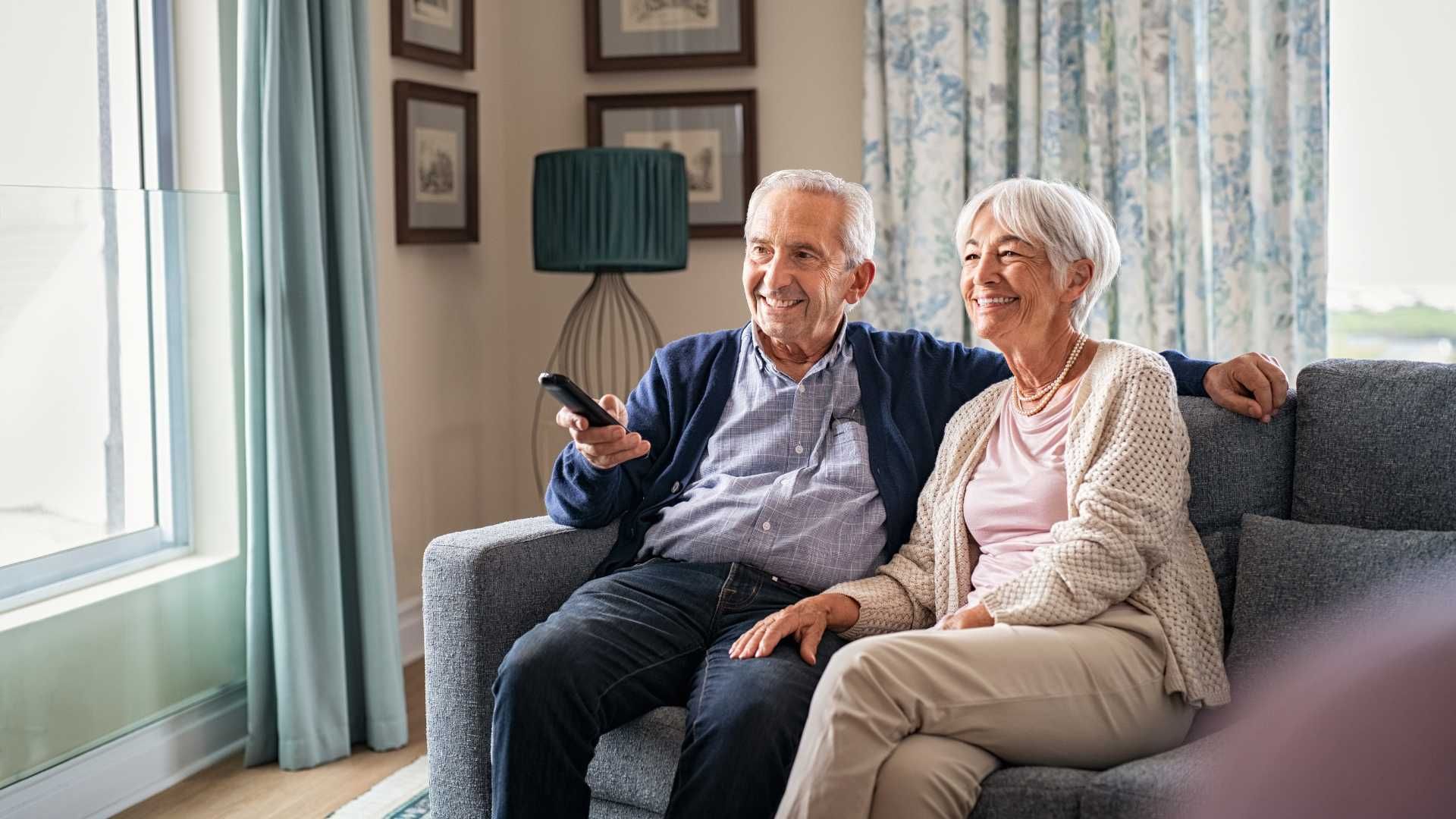 An elderly couple is sitting on a couch watching television.