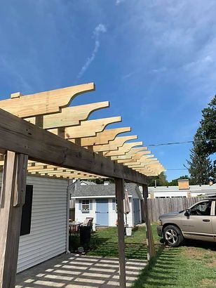 A car is parked under a wooden pergola in front of a house.