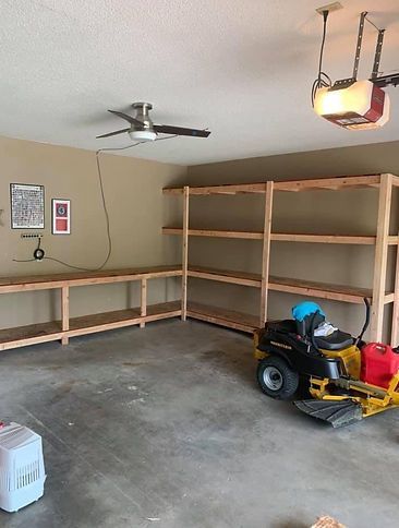 A lawn mower is parked in a garage with shelves and a ceiling fan.