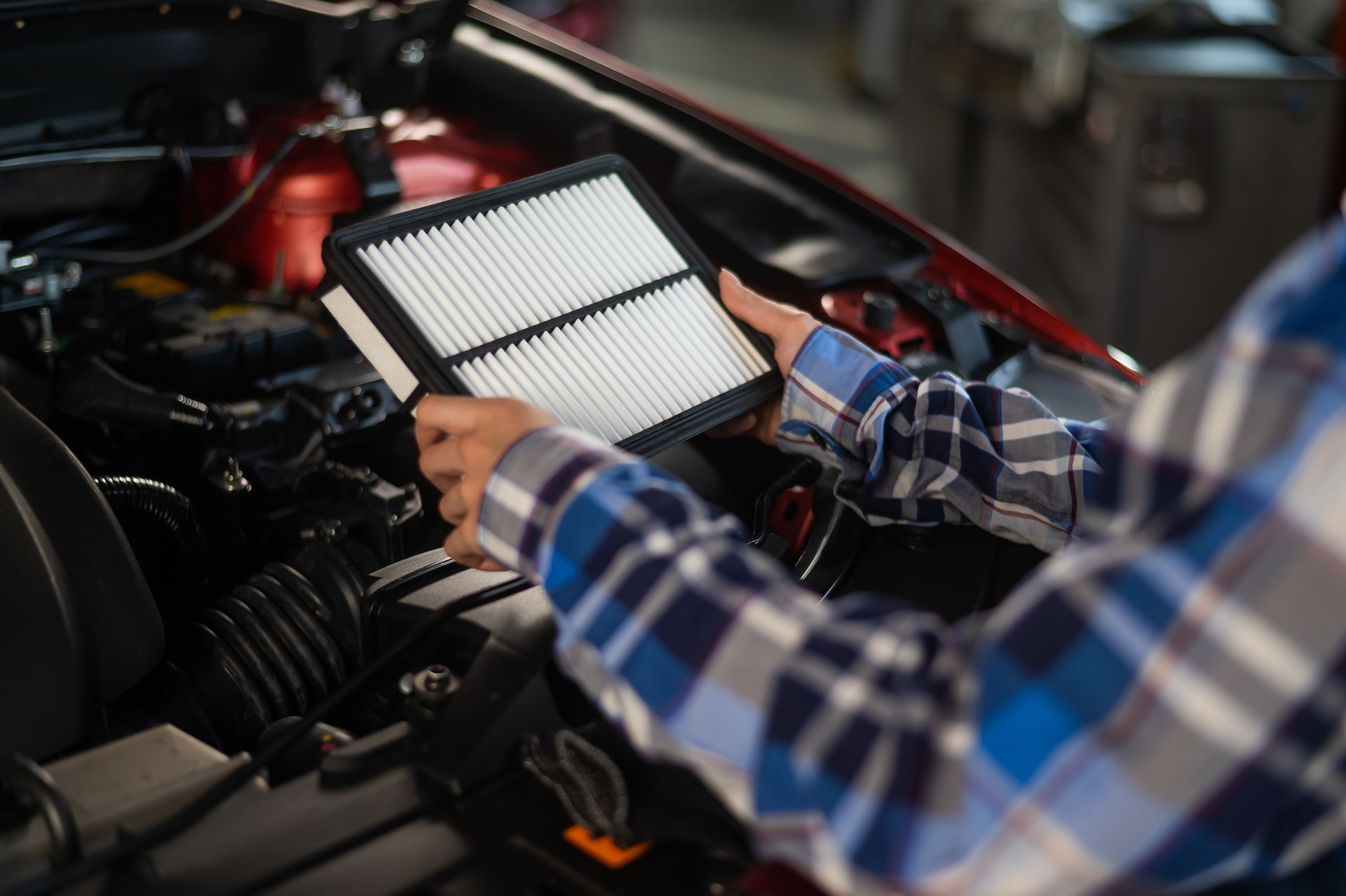 Person installing a white air filter in a car engine bay, wearing a blue plaid shirt