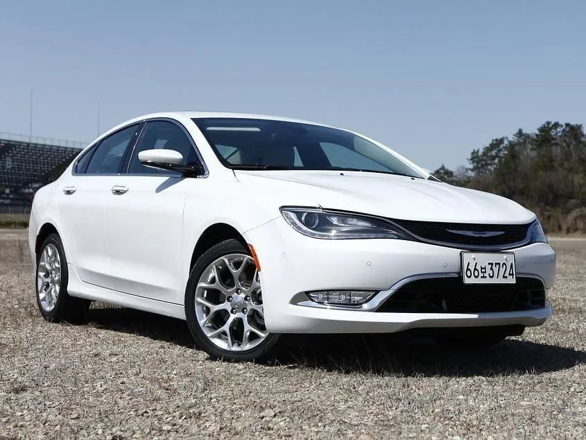 White sedan parked on gravel under a clear sky, front view with chrome grille and alloy wheels