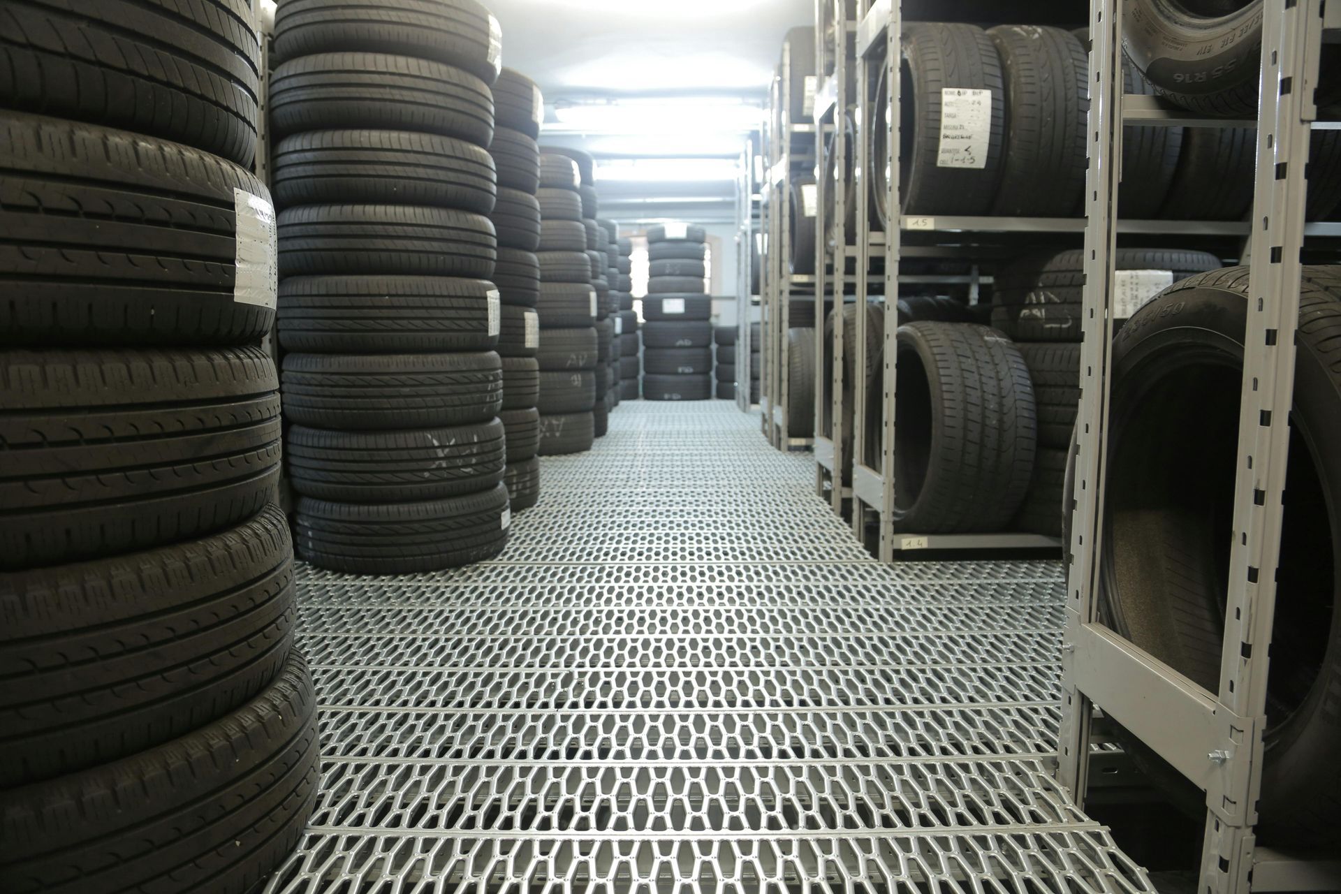 Rows of stacked car tires in a warehouse aisle with metal shelving and a grated floor