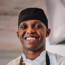 Un chef portant un chapeau noir et un tablier sourit.