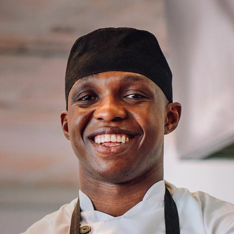 Un chef portant un chapeau noir et un tablier sourit.
