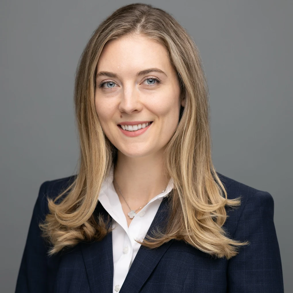 Woman with long blonde hair smiles, wearing a navy blazer over a white shirt, against a gray backdrop.