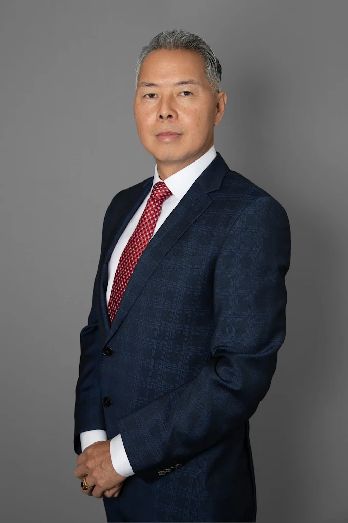 A middle-aged Asian man in a navy suit with a red tie, smiling slightly. Gray background.